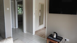 A hotel room with a television mounted on the wall, a small table with a kettle, and a view of a balcony with greenery.