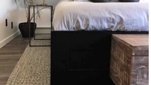 Een slaapkamer met een bed, nachtkastje, lamp, potplant en een houten dressoir.