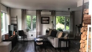 Een moderne woonkamer met een grijze bank, een salontafel en een televisie op een standaard. Er zijn grote ramen met gordijnen, een open haard en een stenen muur.