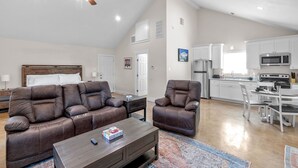 Un vaste espace de vie doté d’un haut plafond, d’un ventilateur de plafond, d’une grande fenêtre et d’une cuisine avec des armoires blanches et des appareils électroménagers modernes.
