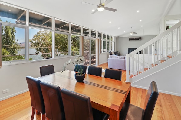 A spacious dining area with a wooden table and chairs, large windows offering a view of the outdoors, and a staircase leading to an upper level.