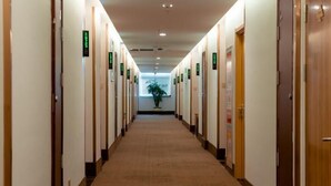 A hotel corridor with doors marked by green signs, carpeted flooring, and a potted plant at the end. - Greentree Inn Jiangsu Suzhou Zhangjiagang Tangshi Town Yangzi Road Express Hotel (Suzhou)