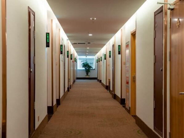 A hotel corridor with doors marked by green signs, carpeted flooring, and a potted plant at the end. - Greentree Inn Jiangsu Suzhou Zhangjiagang Tangshi Town Yangzi Road Express Hotel (Suzhou)