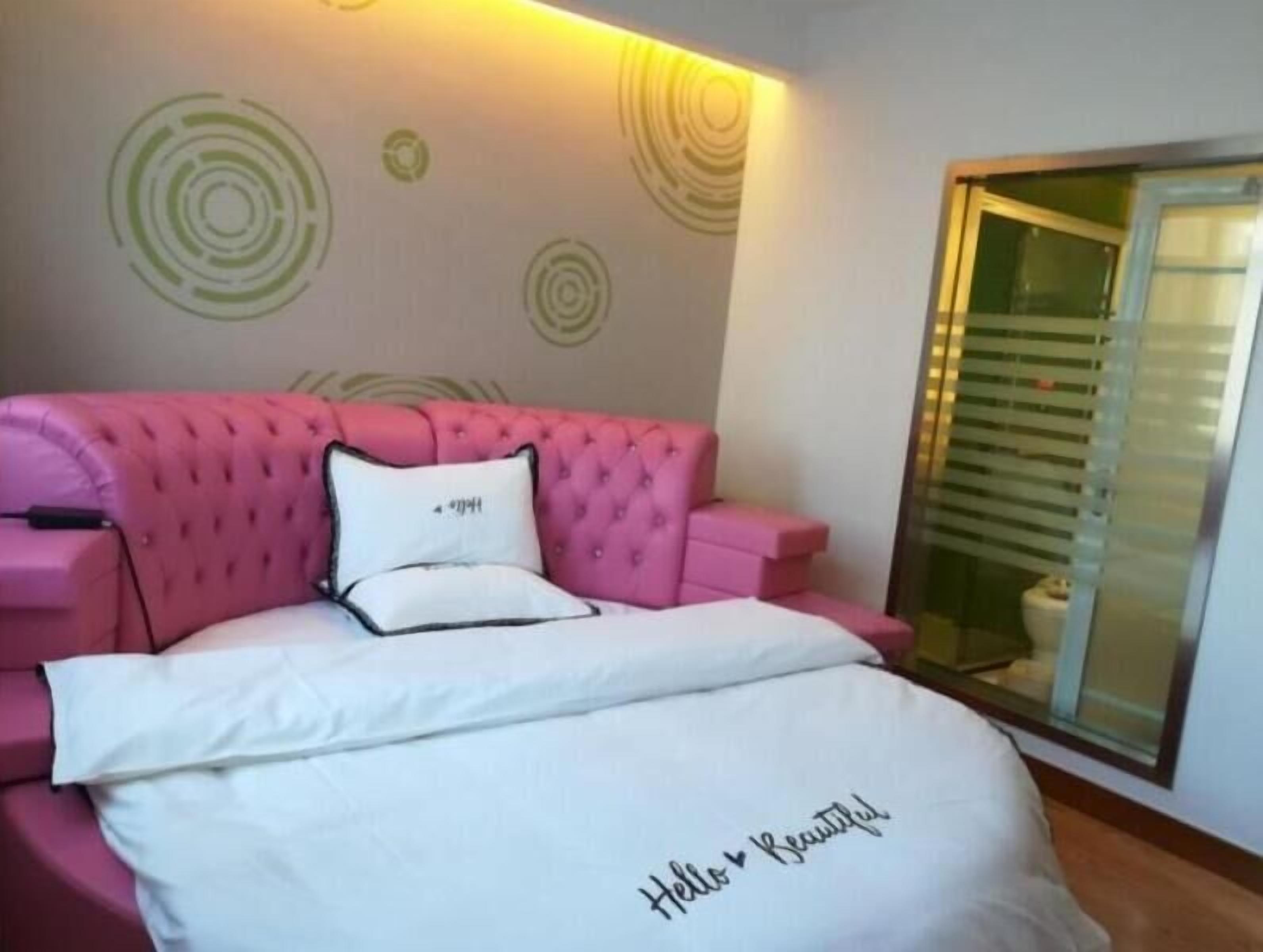 A bedroom with a pink tufted sofa bed, a white bedspread with "Hello Beautiful" embroidered, and a wall with circular patterns.