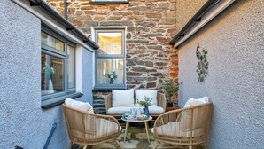 Un espace de détente extérieur accueillant, avec des fauteuils en osier et un canapé blanc, un mur en pierre et une fenêtre agrémentée de rideaux.