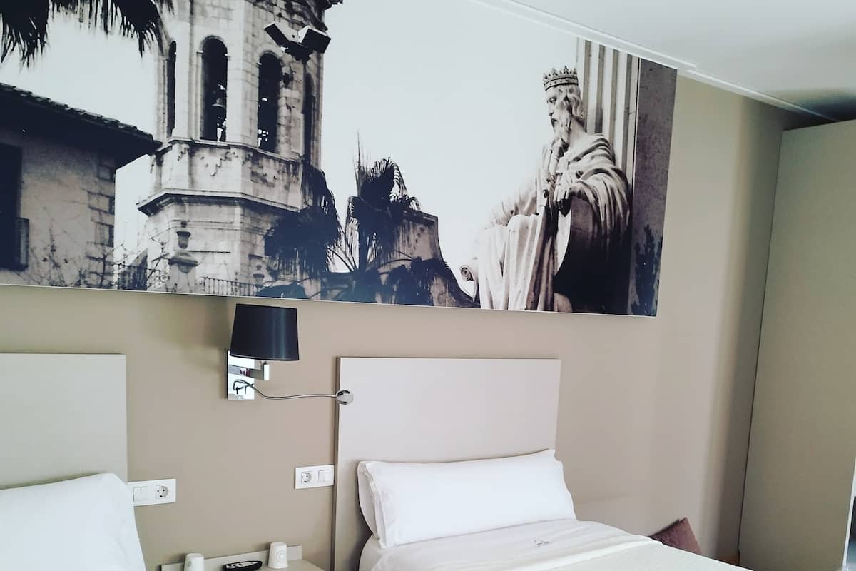 A hotel room with two beds, a nightstand, a wall-mounted TV, and a large framed picture of a historical building.