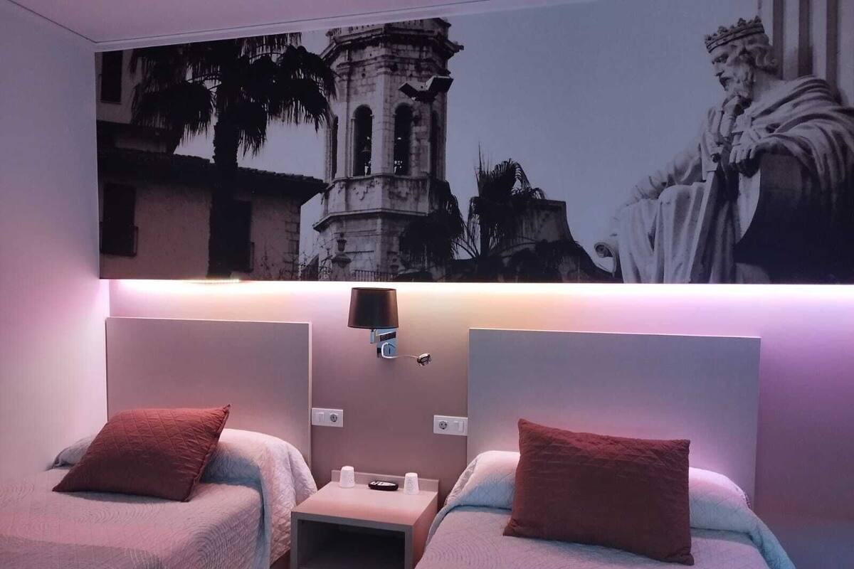 A hotel room with two beds, a nightstand, a wall-mounted lamp, and a large framed picture of a historical building.