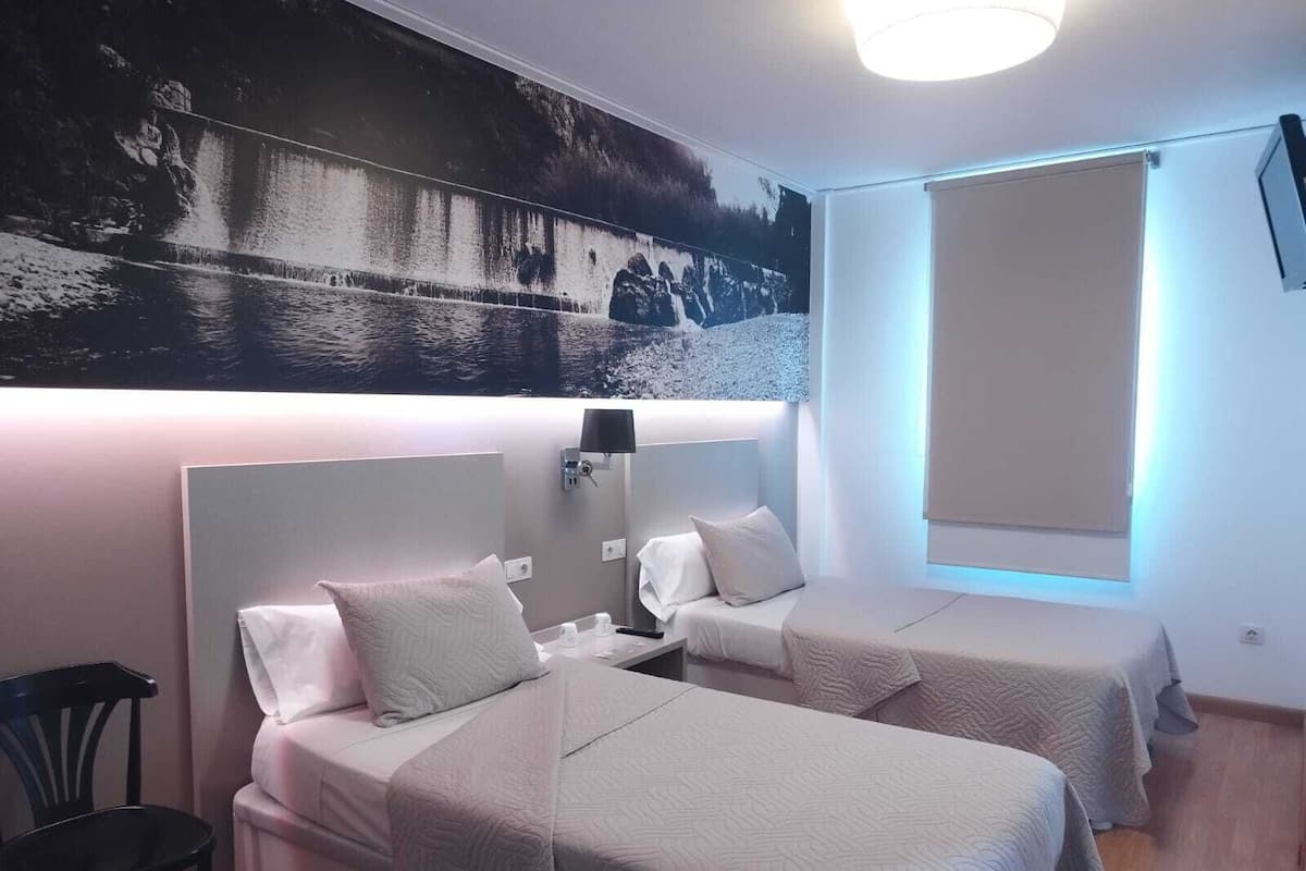 A hotel room with two beds, a chair, a wall-mounted TV, and a large window.