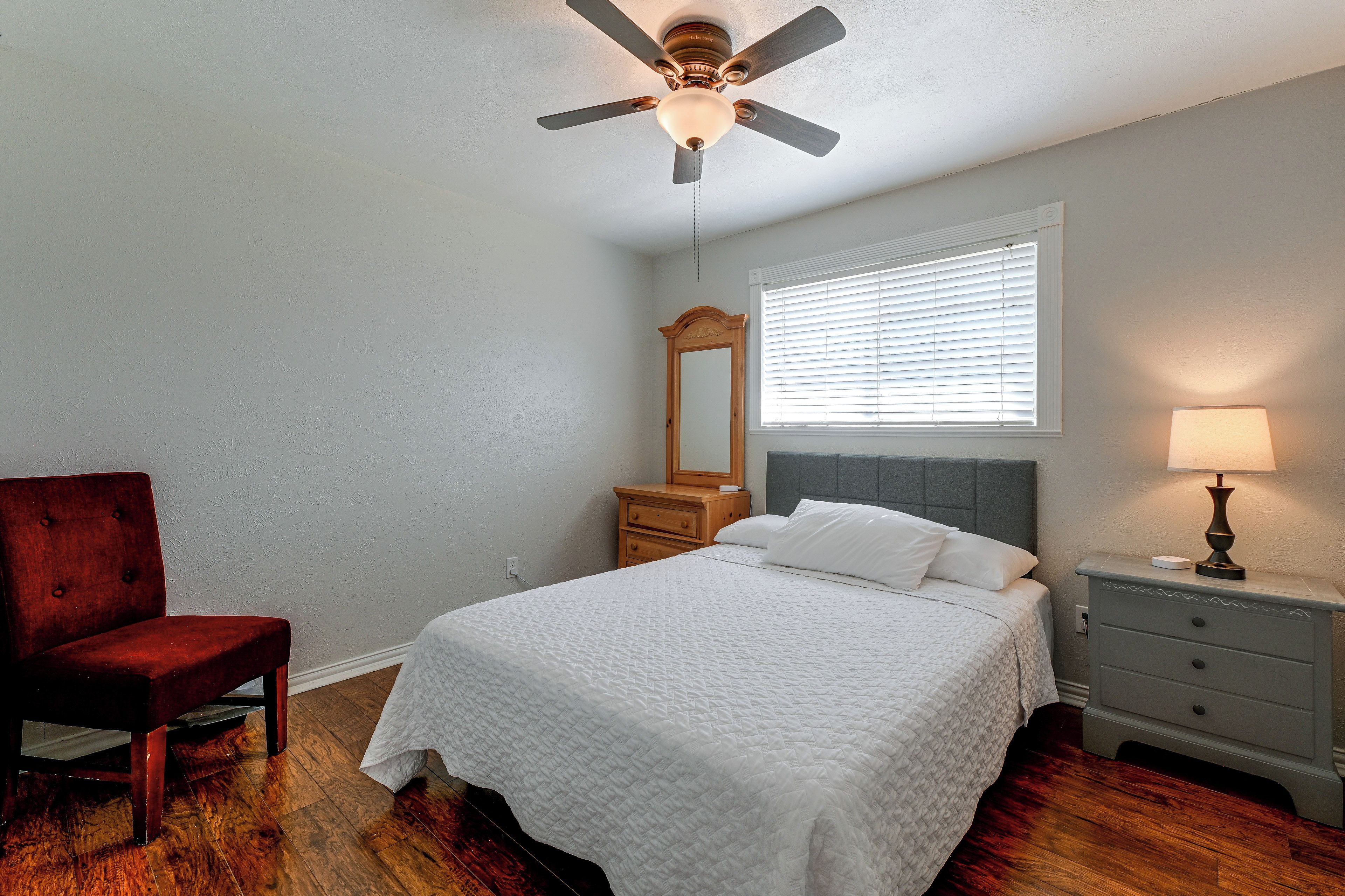 A bedroom with a bed, a chair, a nightstand, a lamp, a mirror, and a ceiling fan.