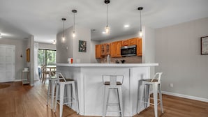 Una cocina moderna con armarios de madera, una isla central con taburetes altos e iluminación colgante, y vistas al área de comedor.
