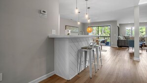 Una cocina moderna con una isla blanca, taburetes de bar, iluminación colgante y vistas al área de estar exterior.