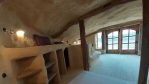 Ein rustikales Dachzimmer mit schragem Dach, Holzbalken und einem großen Fenster mit Blick ins Freie.