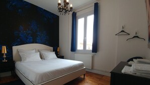 Un dormitorio con cama, candelabro, ventana con cortinas, radiador y una mesa con toallas.