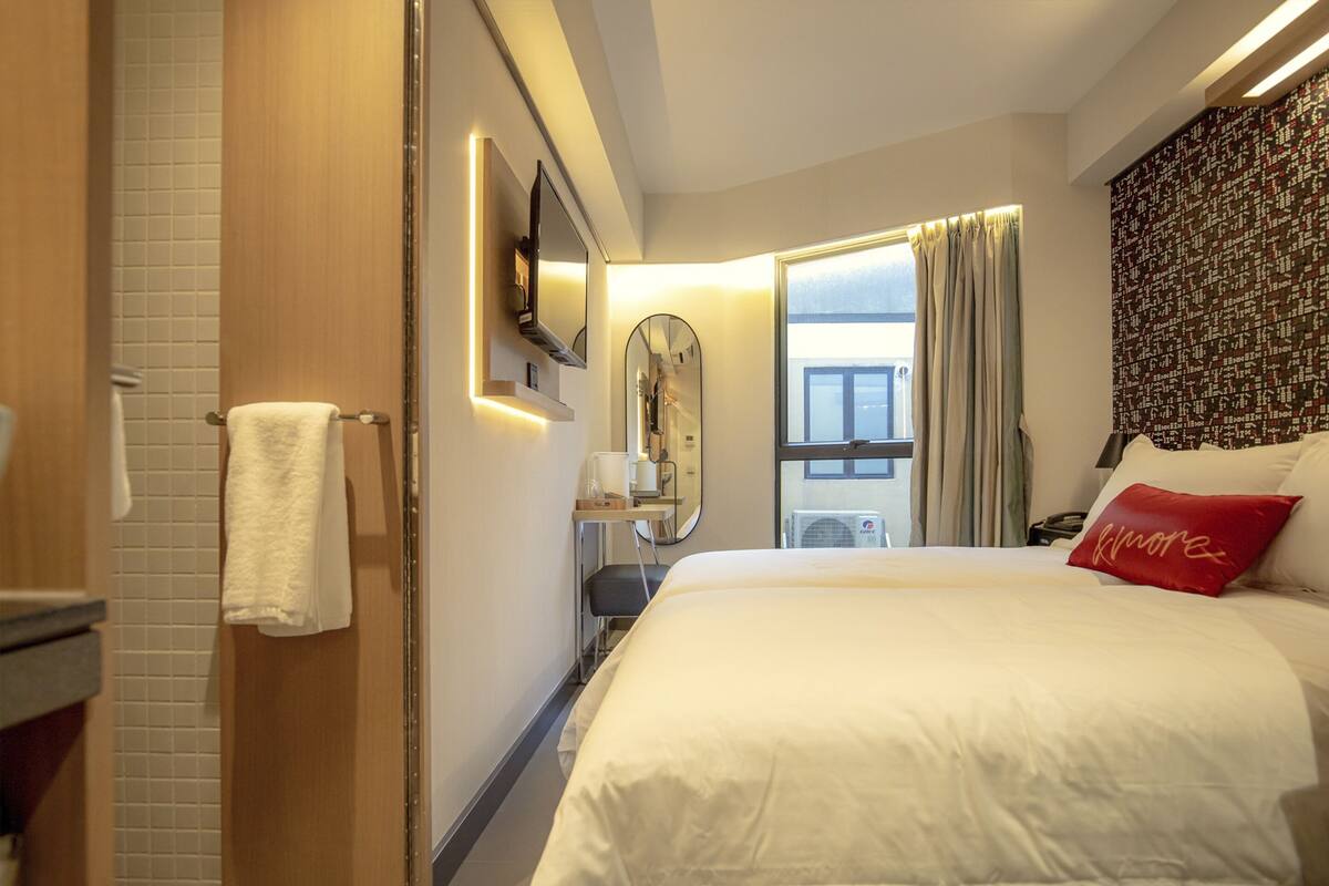 A hotel room with a large bed, a mirror, a bedside table, and a window with curtains.