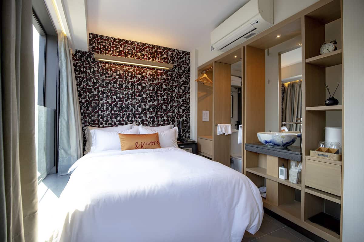 A hotel room with a large bed, a bedside table, a wardrobe, a bathroom with a sink and mirror, and a patterned wall.