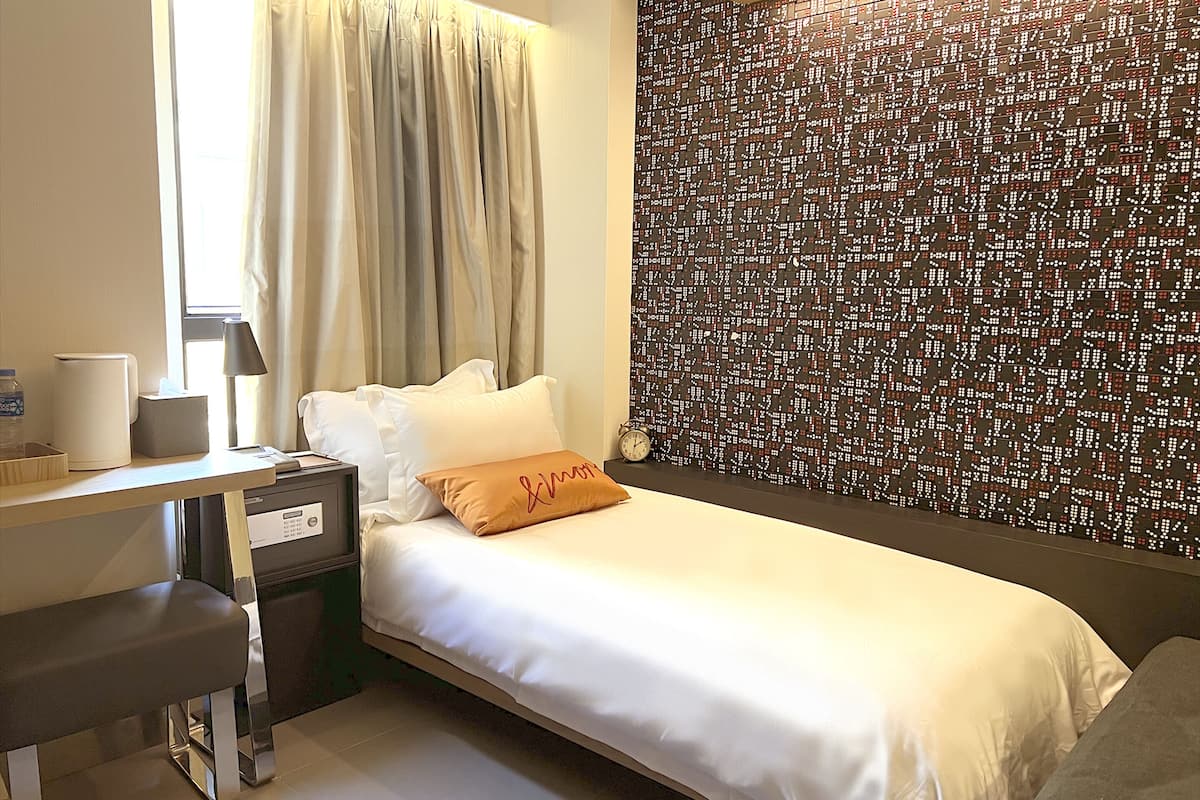 A hotel room with a bed, a desk, a chair, a nightstand, a lamp, and a wall with a patterned design.