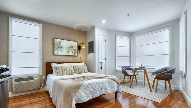 A bedroom with a bed, a desk, two chairs, a window, and a wall-mounted lamp.