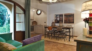 A cozy dining area with a wooden table and chairs, a green sofa with cushions, and a large window with a view of greenery. - San Ruffino Apartments (Casciana Terme Lari)