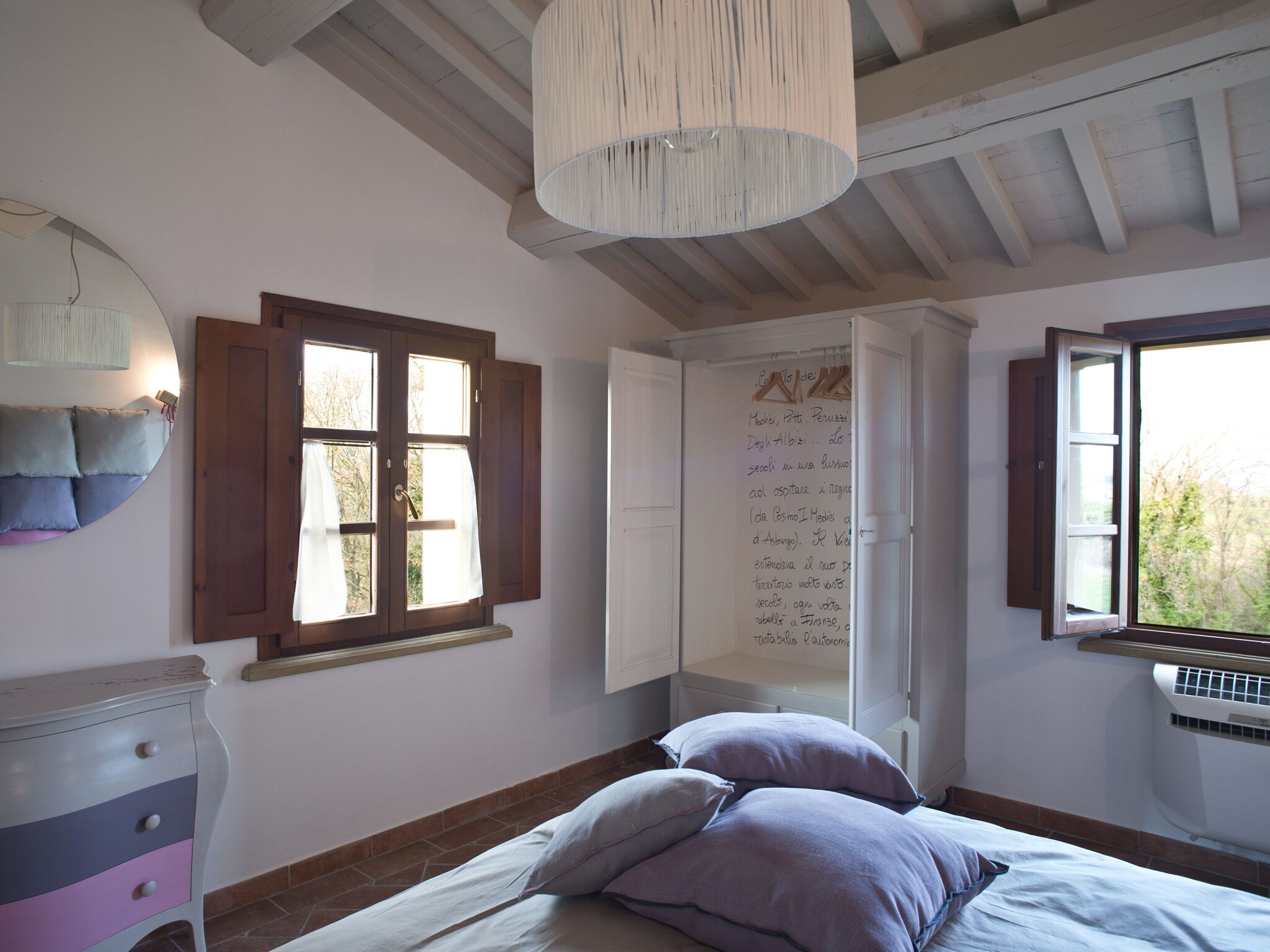A bedroom with a bed, a dresser, a window with open shutters, and a wall with handwritten notes.