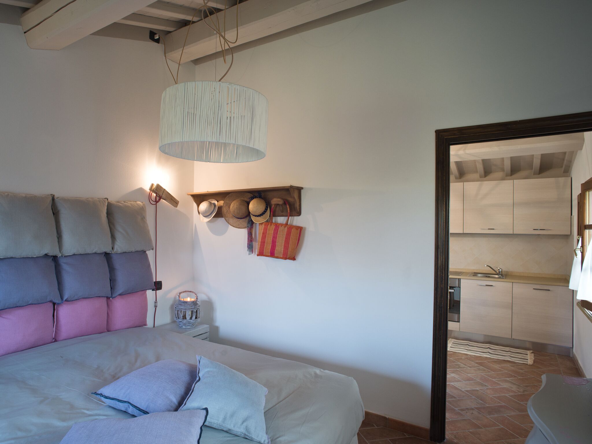 Una camera da letto con un letto, cuscini, una lampada da comodino, una mensola a parete con cappelli e borse, e vista sulla cucina.
