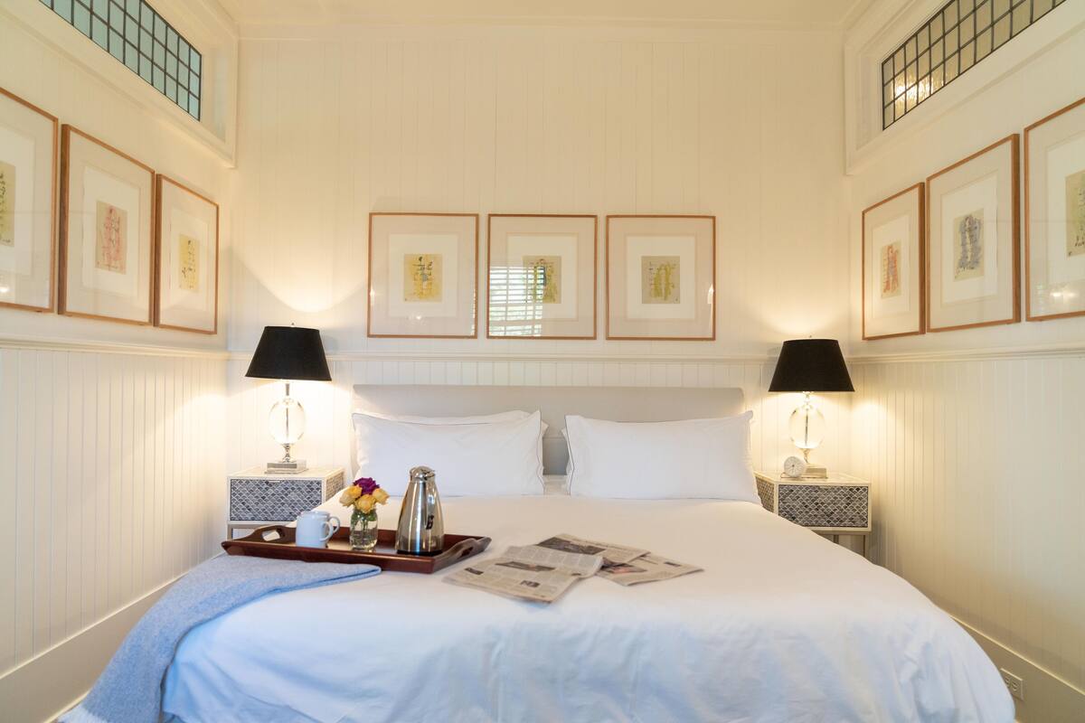 A neatly made bed with white linens, two bedside lamps, a tray with a teapot, a vase with flowers, and framed pictures on the wall.