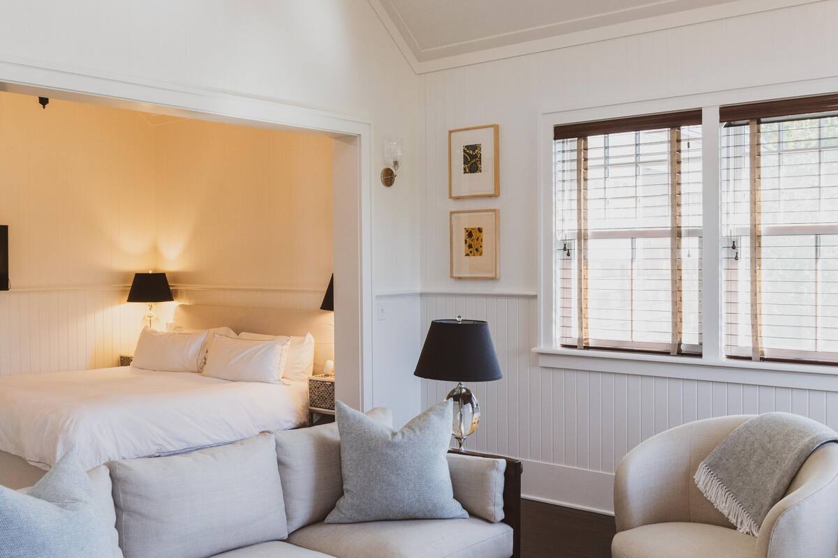 A room with a bed, a sofa, a chair, a coffee table, and a window with blinds.