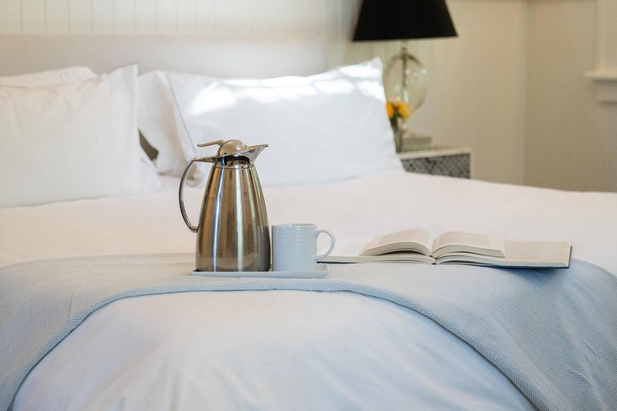 A neatly made bed with a metal coffee pot, a book, and a cup on the bedside table.