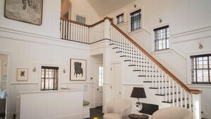 A spacious living room with a staircase, a fireplace, and a coffee table with books and flowers. - The Reform Club (Amagansett)