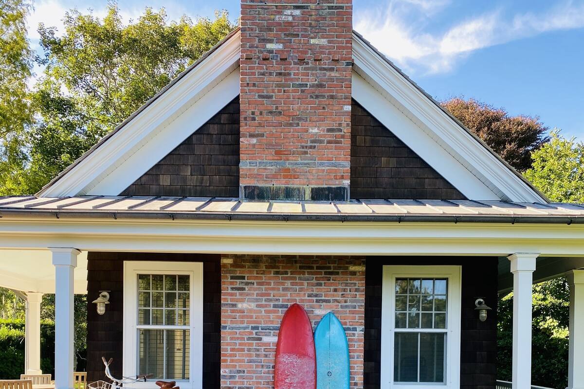A house with a brick chimney, two windows, and a porch with a bicycle and surfboards.