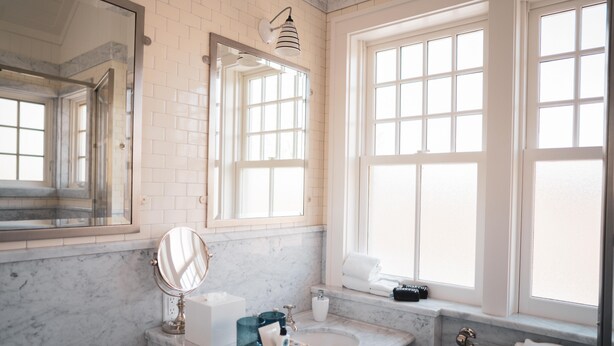 A bathroom with a marble sink, a large mirror, a window, and a towel rack. - The Reform Club (Amagansett)