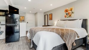Ein Schlafzimmer mit Bett, Nachttischen, einem kleinen Tisch, einem schwarzen Kühlschrank, einer weißen Tür und einem Bild an der Wand.