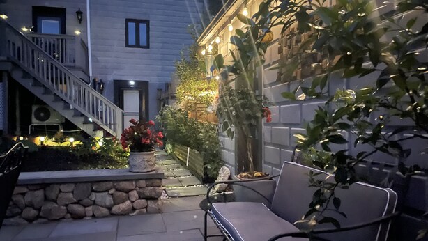 Ein Innenhof mit einer Steinmauer, einer Bank, einem Blumentopf mit roten Blüten und einer Treppe, die zu einer oberen Ebene führt.