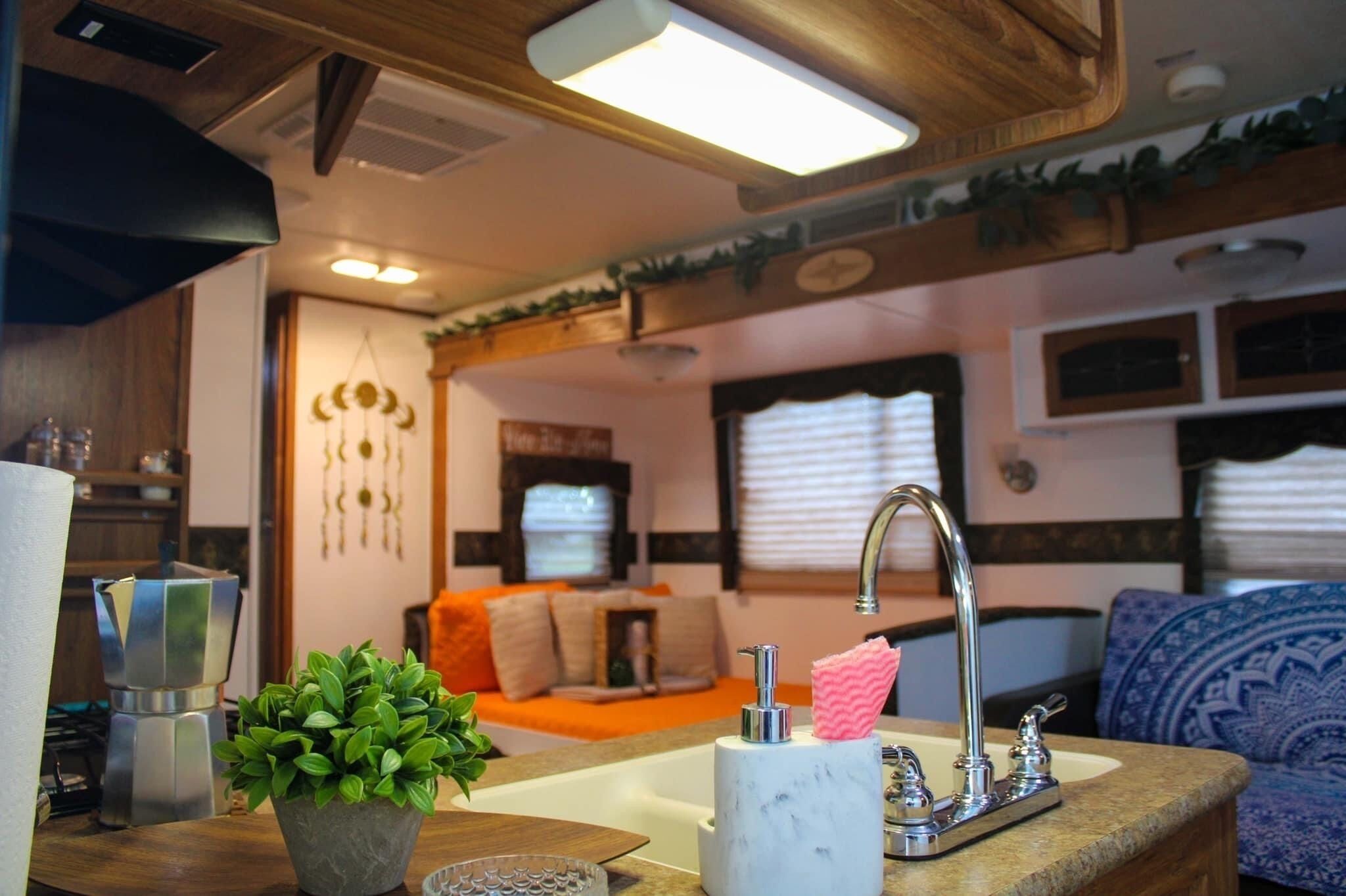 A compact RV interior with a kitchenette, a seating area with cushions, and a window with blinds.