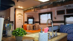 Un intérieur de camping-car compact comprenant un coin cuisine, un espace salon avec des coussins et une fenêtre équipée de stores.