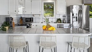 Una cocina moderna con armarios blancos, un refrigerador de acero inoxidable, una isla central con un frutero y tres sillas de mimbre.