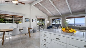 Una cocina amplia con una isla grande, muebles blancos y un ventilador de techo. Hay un comedor con una mesa y sillas de madera, y vistas al océano a través de grandes ventanales.