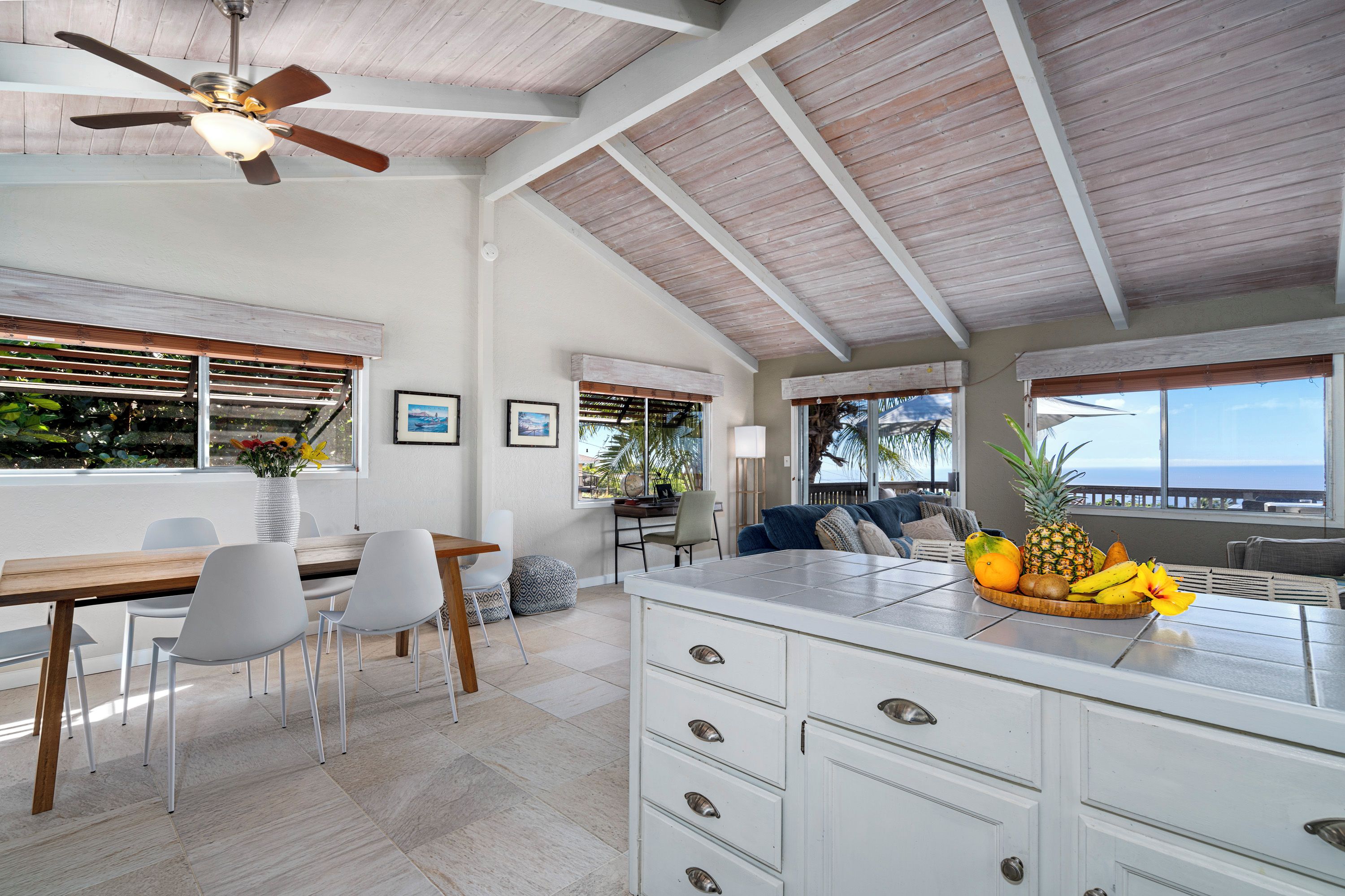A spacious kitchen with a large island, white cabinetry, and a ceiling fan. There is a dining area with a wooden table and chairs, and a view of the ocean through large windows.