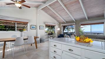 A spacious kitchen with a large island, white cabinetry, and a ceiling fan. There is a dining area with a wooden table and chairs, and a view of the ocean through large windows.