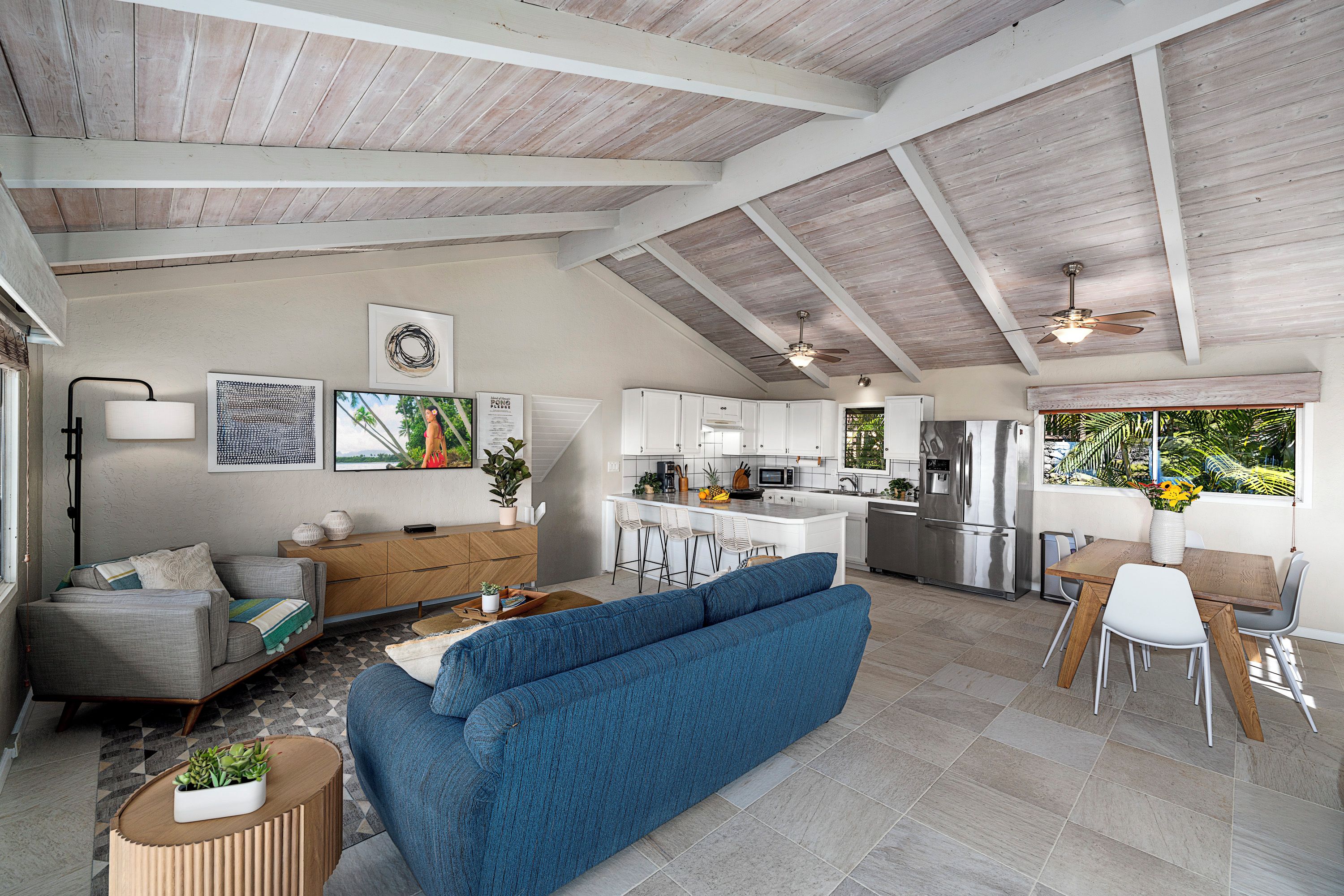 A spacious living area with a blue sofa, a wooden dining table with chairs, a kitchen with white cabinets, and a ceiling fan.