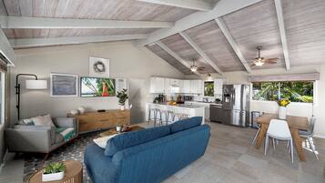 A spacious living area with a blue sofa, a wooden dining table with chairs, a kitchen with white cabinets, and a ceiling fan.