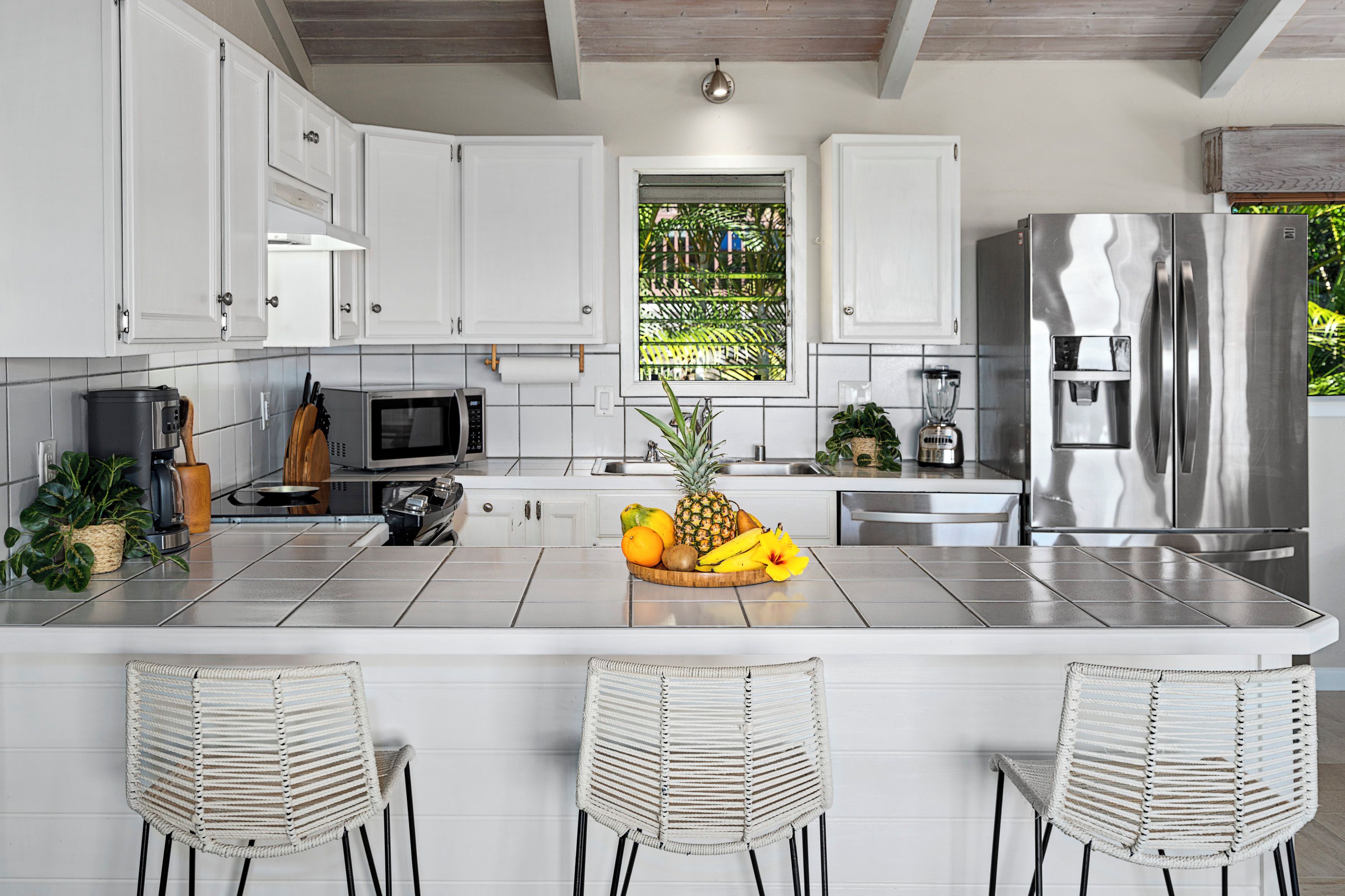A modern kitchen with white cabinets, a stainless steel refrigerator, a central island with a fruit bowl, and three wicker chairs.