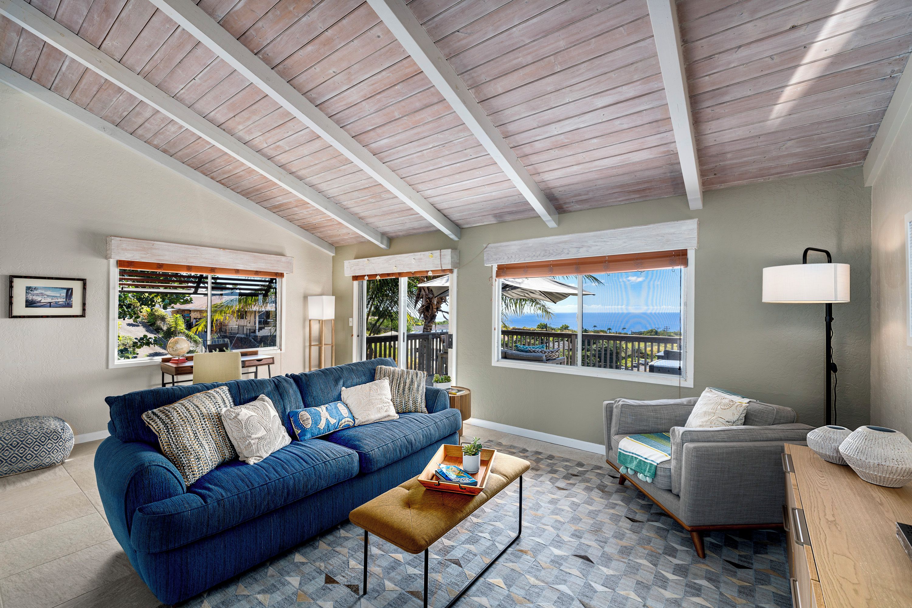 A living room with a blue sofa, a grey armchair, a wooden coffee table, and a view of the ocean through large windows.