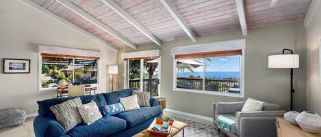 A living room with a blue sofa, a grey armchair, a wooden coffee table, and a view of the ocean through large windows.