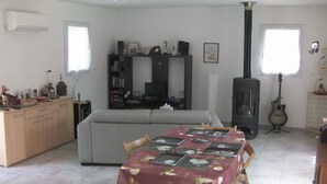 Ein Wohnzimmer mit einer Couch, einem Esstisch mit Blumenmuster-Tischdecke, einem TV-Schrank mit Lautsprechern, einer Gitarre und einem Kamin.