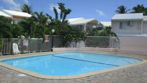 A swimming pool surrounded by a white fence, with palm trees and a building in the background. - Mezzanine À LA Marina (Saint-François)