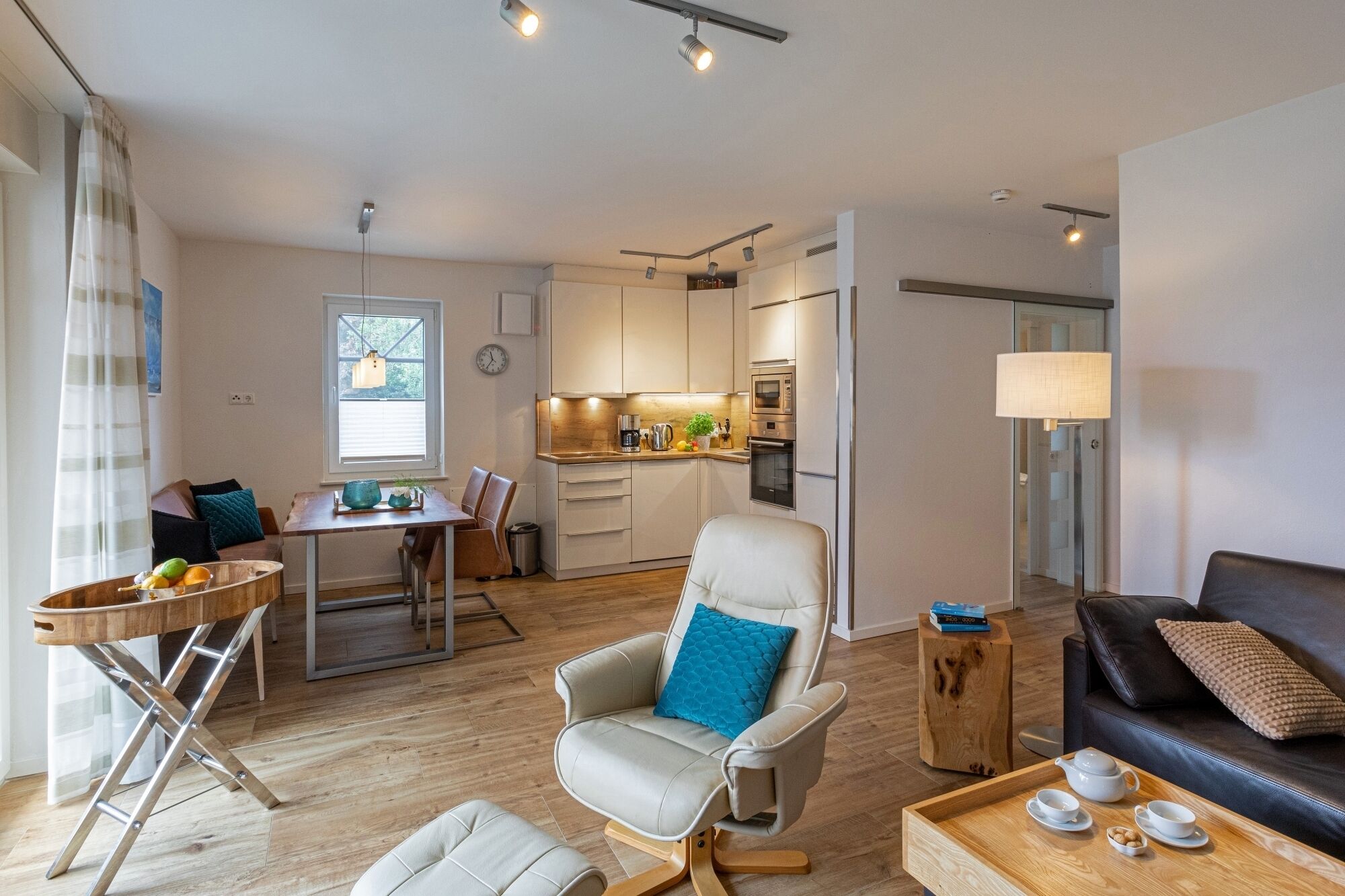 Ein modernes Wohnzimmer mit Ledersofa, Couchtisch mit Teeservice, Essbereich mit Holztisch und Stühlen sowie Küche mit weißen Schränken und Edelstahlgeräten.