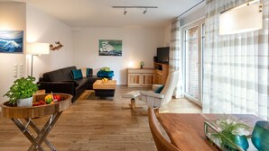 Una sala de estar moderna con un sofá negro, un sillón blanco, una mesa de centro de madera y un televisor sobre un mueble de madera.