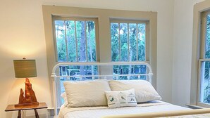 Un dormitorio con estructura de cama de metal blanco, suelo de madera y vistas a árboles a través de amplios ventanales.