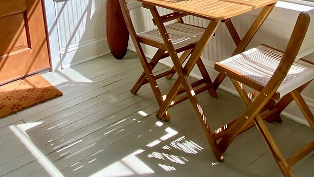 Una habitación iluminada por el sol con una mesa y sillas de madera, una planta en maceta y un ventanal con vistas a árboles.