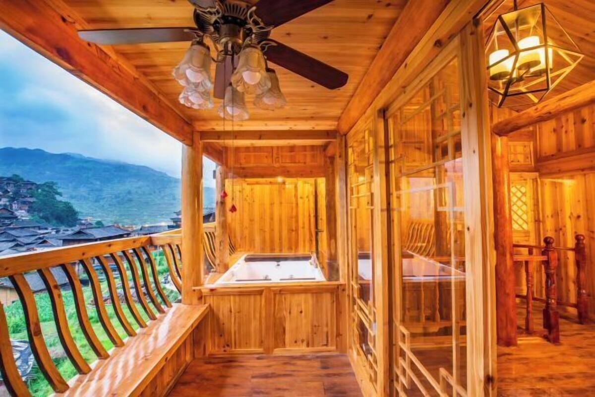 Una cabaña de madera con balcón, ventilador de techo y un ventanal grande con vista a un paisaje montañoso.